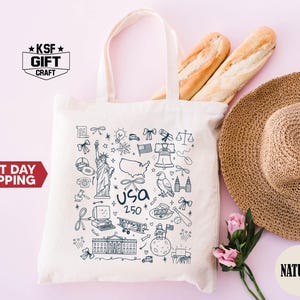 May include: Cream-colored tote bag with a blue USA-themed doodle design. The bag features illustrations of the Statue of Liberty, a map, and other patriotic symbols. A straw hat, two baguettes, and pink flowers are next to the bag.