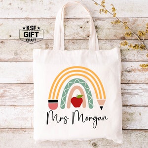 May include: A white tote bag with a rainbow design, an apple, and two pencils. The text "Mrs. Morgan" is printed below the rainbow.