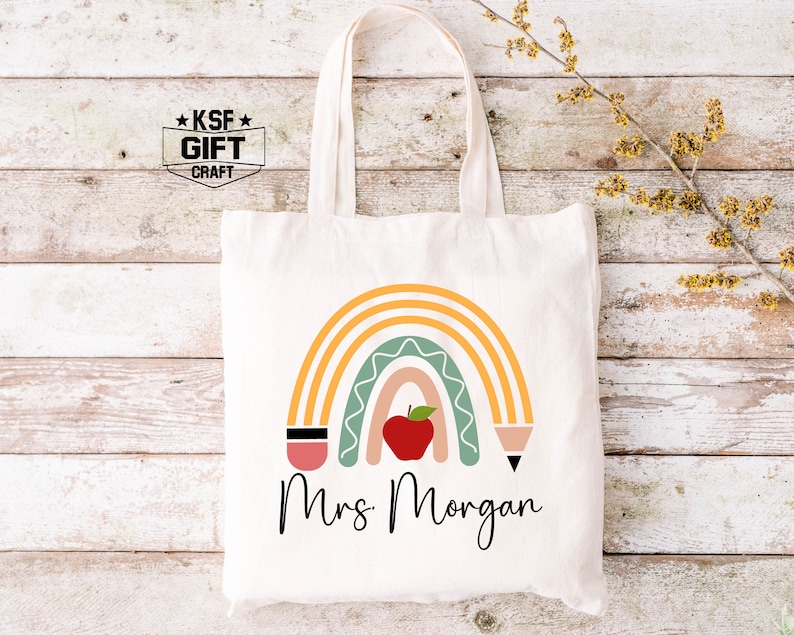 May include: A white tote bag with a rainbow design, an apple, and two pencils. The text "Mrs. Morgan" is printed below the rainbow.