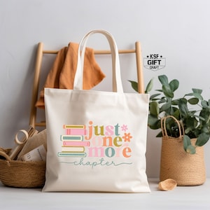Op de afbeelding: Een witte canvas tote bag met een kleurrijk grafisch ontwerp met de tekst "Just one more chapter" met een stapel boeken. Het grafische ontwerp is in roze, blauw, groen en geel.