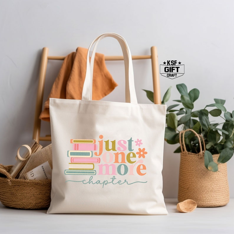 Op de afbeelding: Een witte canvas tote bag met een kleurrijk grafisch ontwerp met de tekst "Just one more chapter" met een stapel boeken. Het grafische ontwerp is in roze, blauw, groen en geel.