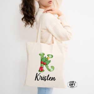 May include: A white canvas tote bag featuring a green holly leaf letter K with a red berry and gold star embellishment. The name Kristen is printed in black below the letter.