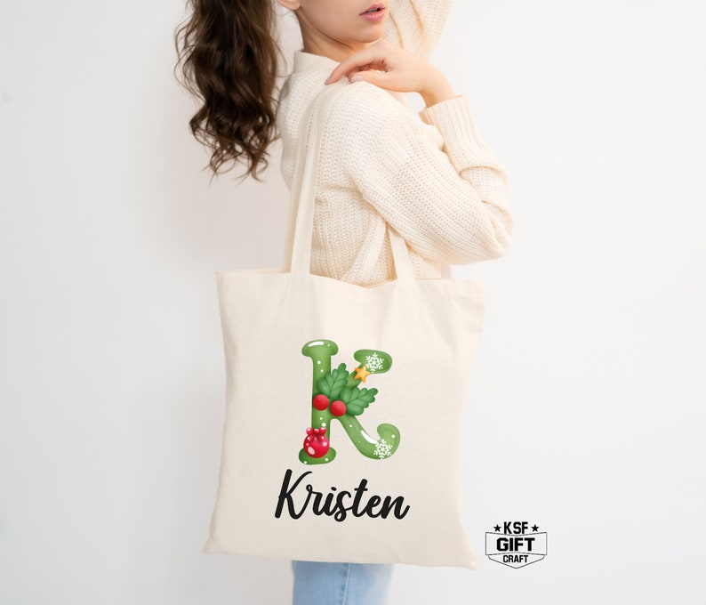 May include: A white canvas tote bag featuring a green holly leaf letter K with a red berry and gold star embellishment. The name Kristen is printed in black below the letter.