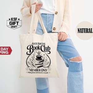 Op de afbeelding: Cr&egrave;mekleurige draagtas met de tekst "ANTI SOCIAL Book Club" in een decoratief lettertype. Het ontwerp bevat een stapel boeken, een kopje koffie en de zin "MEMBER ONLY: Making friends with characters, not people."