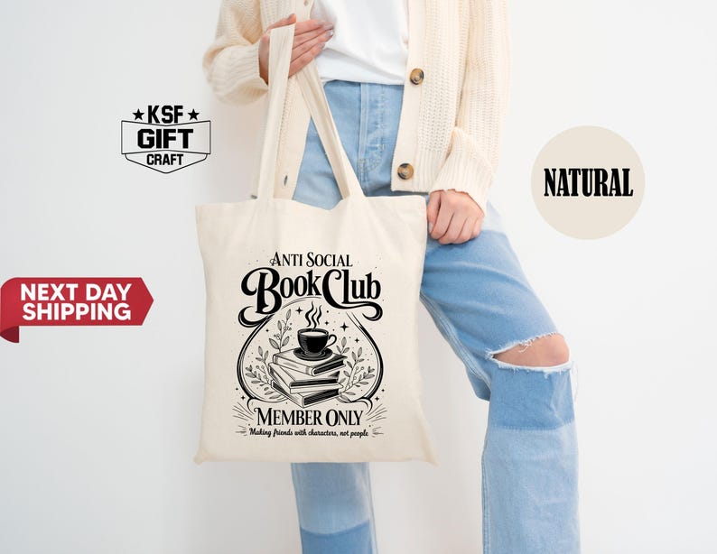 Op de afbeelding: Cr&egrave;mekleurige draagtas met de tekst "ANTI SOCIAL Book Club" in een decoratief lettertype. Het ontwerp bevat een stapel boeken, een kopje koffie en de zin "MEMBER ONLY: Making friends with characters, not people."