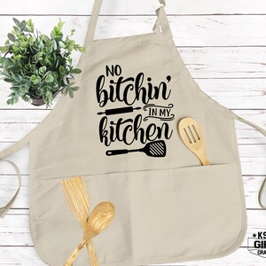 Może przedstawiać: Beżowy fartuch kuchenny z dwoma kieszeniami i czarnym nadrukiem z napisem "No bitchin' in my kitchen" z wałkiem i szpatułką.