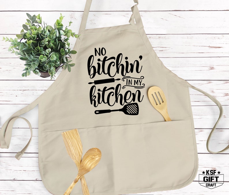 Może przedstawiać: Beżowy fartuch kuchenny z dwoma kieszeniami i czarnym nadrukiem z napisem "No bitchin' in my kitchen" z wałkiem i szpatułką.