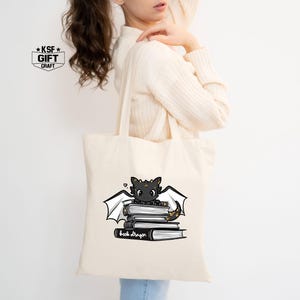Pu&ograve; includere: Una borsa tote bianca con un'illustrazione in bianco e nero di un drago seduto su una pila di libri. Il testo "Book Dragon" &egrave; stampato sotto il drago.