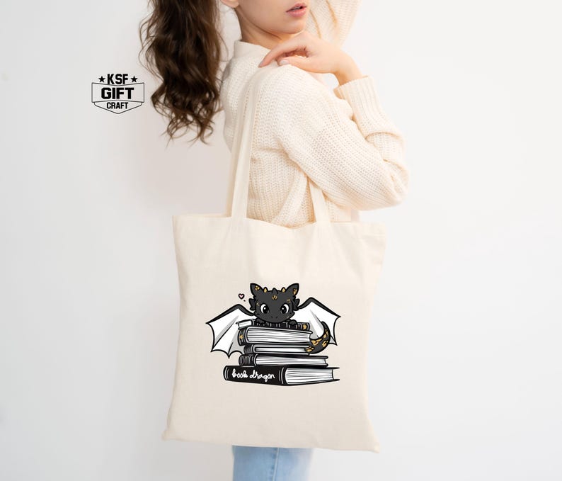 Pu&ograve; includere: Una borsa tote bianca con un'illustrazione in bianco e nero di un drago seduto su una pila di libri. Il testo "Book Dragon" &egrave; stampato sotto il drago.