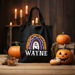 May include: Black canvas tote bag with a rainbow design featuring a ghost wearing sunglasses. The rainbow is made up of orange, purple, and white stripes with white stars and bats. The text "WAYNE" is printed below the rainbow.