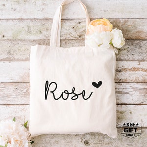 May include: A white canvas tote bag with the name "Rose" written in black cursive script with a small black heart next to it. The bag is on a white wood background.
