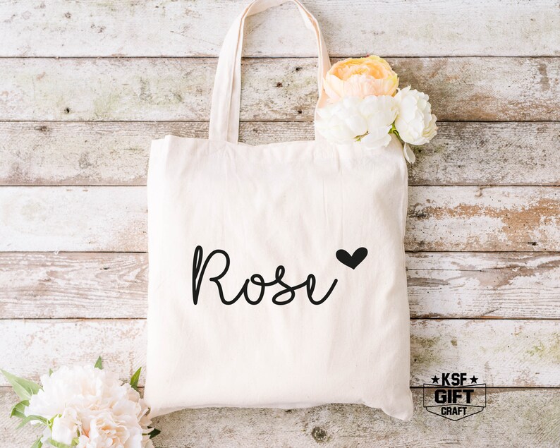 May include: A white canvas tote bag with the name "Rose" written in black cursive script with a small black heart next to it. The bag is on a white wood background.