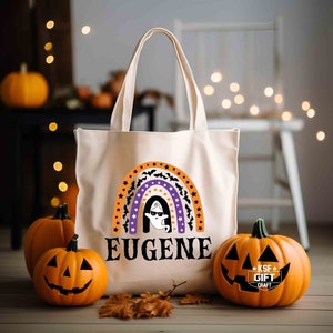 May include: A white canvas tote bag with a rainbow design featuring orange, purple and white stripes, black bats and white stars. The rainbow has a ghost inside and the text "EUGENE" below it. The bag is sitting on a wooden surface with two orange pumpkins.