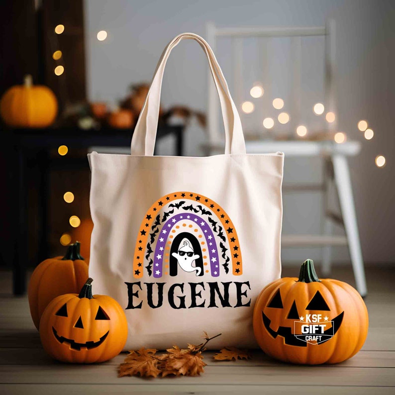 May include: A white canvas tote bag with a rainbow design featuring orange, purple and white stripes, black bats and white stars. The rainbow has a ghost inside and the text "EUGENE" below it. The bag is sitting on a wooden surface with two orange pumpkins.