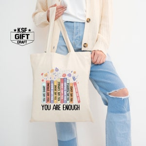 May include: A beige tote bag with a design of colorful books stacked on top of each other. The books have positive affirmations printed on them, such as "You are smart", "You are strong", "You are kind", and "You are loved". The text "You are enough" is printed at the bottom of the bag.