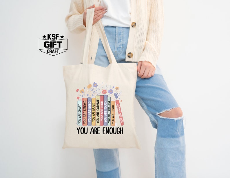 May include: A beige tote bag with a design of colorful books stacked on top of each other. The books have positive affirmations printed on them, such as "You are smart", "You are strong", "You are kind", and "You are loved". The text "You are enough" is printed at the bottom of the bag.