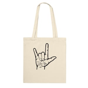 Puede incluir: Una bolsa de tela beige con una ilustración negra del símbolo del lenguaje de señas americano para "Te amo". El texto "Te amo" está escrito debajo del signo de la mano.