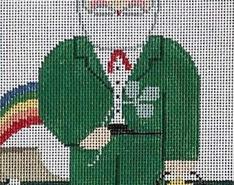 May include: A needlepoint canvas featuring a leprechaun wearing a green coat and hat with a red band. The figure has a white beard, holds a black pot of gold, and stands in a green field with white sheep, a rainbow, and a shamrock emblem.