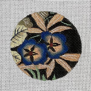 Könnte beinhalten: Eine kreisförmige Stickleinwand mit einem Blumenmuster, das zwei markante blaue Blüten mit dunklen Mitten zeigt, umgeben von schwarzen, beigen und grünen Blattmustern auf einem weißen Gitterhintergrund, was ein detailliertes Textilkunstwerk ergibt.