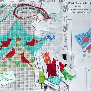 May include: Needlepoint canvas featuring a star design with red birds and a gold star. The canvas is accompanied by various threads, including green, red, and white, and a printed pattern guide. The text on the canvas reads "Christmas Star with Red Birds".