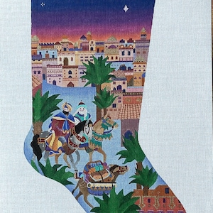 Puede incluir: Un lienzo de punto de aguja con forma de calcetín navideño que muestra una ciudad en el desierto al atardecer. El diseño incluye tres figuras montadas en camellos, palmeras y un paisaje urbano bajo un cielo azul y morado. Las iniciales NAN están abajo.