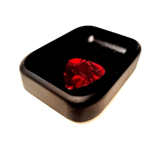 May include: A black rectangular guitar pick holder with a red guitar pick inside. The pick has a textured surface. The holder has a smooth, glossy finish.