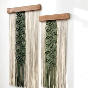 May include: Two macrame wall hangings with a green leaf pattern and a natural beige color. The hangings are attached to wooden dowels.