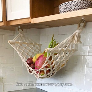 May include: A white macrame hammock hanging from a cabinet, filled with fresh fruit, including a dragon fruit, bananas, and other tropical fruits. The hammock is made of natural cotton rope and has a wooden dowel at the top.