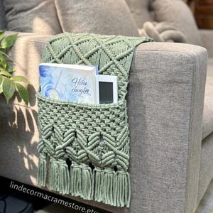 May include: A sage green macrame sofa armrest organizer with a pocket, holding a book and a small electronic device. The organizer features a geometric pattern and fringe detailing. The text "Hieu chuyen" is visible on the book. The Etsy shop name is visible at the bottom.
