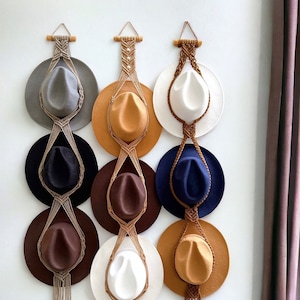 May include: A wall-mounted hat rack made of macrame with six hats hanging on it. The hats are in various colours, including brown, white, grey, and blue. The rack is made of natural fibres and has a rustic look. The rack is hanging on a white wall, and there is a brown leather sofa in the foreground.