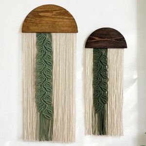 May include: Two macrame wall hangings with a green leaf pattern and a wooden half-circle top. The larger hanging has a light brown wood top and the smaller hanging has a dark brown wood top.