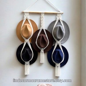 May include: A macrame hat rack with four hats hanging on it. The rack is made of white cord and has a wooden dowel at the top. The hats are brown, gray, black, and blue. The rack is hanging on a white wall. The text "lindecormacramestore.etsy.com" is visible at the bottom of the image. A straw hat with a pink ribbon is sitting on a wooden table.
