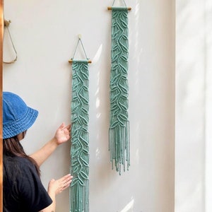 Puede incluir: Dos tapices de macramé de color verde claro con una barra de madera en la parte superior. Los tapices están hechos con un patrón de hojas y tienen una larga franja en la parte inferior. Los tapices se muestran en una pared blanca.