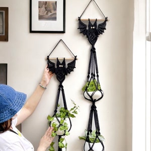 Puede incluir: Dos maceteros de macramé negros con forma de murciélagos con ojos blancos. Cada macetero tiene tres niveles y sostiene una pequeña maceta blanca con una planta verde. Los maceteros están colgados en una pared blanca con cuadros enmarcados.