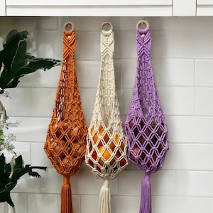 May include: Three macrame hanging produce bags with wooden rings. The bags are orange, white, and purple. Each bag has a tassel at the bottom. The bags are hanging from a white tiled wall. The text "lindecormacramestore.etsy.com" is visible at the bottom of the image.