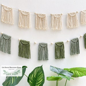 Op de afbeelding: Een macramé-slinger met beige en groene kwastjes hangt aan een witte muur. De slinger is bespannen met houten kralen. De tekst "Lin Decor Macrame Store since 2019" is zichtbaar in de linkerbenedenhoek van de afbeelding.