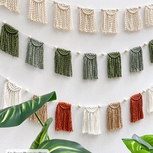 Puede incluir: Una guirnalda de macramé con tres filas de borlas en varios colores, incluyendo crema, verde y naranja. La guirnalda está enhebrada con cuentas de madera y cuelga de una pared blanca. El texto "Lin Decor Macrame Store Since 2019" es visible en la esquina inferior izquierda de la imagen.