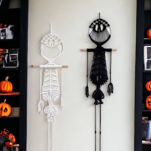 May include: Two macrame wall hangings in the shape of skeletons, one white and one black, hanging on a white wall. The wall hangings are decorated with pumpkins and candles. The text "lindecormacramestore.etsy.com" is visible at the bottom of the image.
