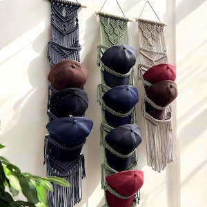 May include: Macrame hat organizers in navy, olive green, and beige, each holding several baseball caps in various colors. The organizers are suspended from wooden dowels, adding a touch of bohemian style to the wall decor.