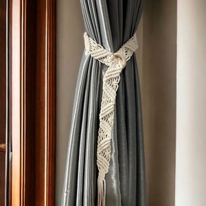 May include: A white macrame curtain tieback with a wooden ring, tied around a gray curtain.