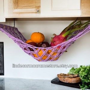 Könnte beinhalten: Lilafarbener Makramee-Hängekorb, der an einem Küchenschrank hängt und Obst wie Orangen, Drachenfrucht und Pflaumen enthält. Der Hängekorb besteht aus geknotetem Seil und hat an jedem Ende einen Holzstab.
