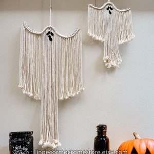 Puede incluir: Dos colgantes de pared de macramé con forma de fantasma con ojos de cuentas negras y un borde con flecos. Un tarro negro, una botella negra y una calabaza naranja tallada con una vela encendida están en una estantería de madera.