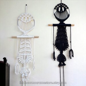May include: Two macrame wall hangings, one white and one black, in the shape of a skeleton with a circular head. The hangings are made with cotton cord and feature a wooden dowel.