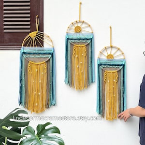 May include: Three macrame wall hangings with a sunburst design in shades of yellow, blue, and green. The hangings are made with natural fibers and feature wooden hoops. The hangings are displayed on a white wall.