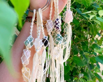 May include: Seven macrame keychains with crystal stones. Each keychain features a small crystal stone, such as rose quartz, amethyst, and clear quartz, encased in a macrame net. The keychains are attached to silver rings and have long white tassels.