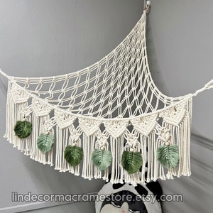 May include: A white macrame hammock with green leaf accents and wooden beads. The hammock is hanging from a hook on a wall.