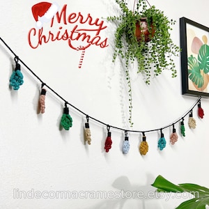 May include: A black string with crocheted lightbulb ornaments in various colors, including red, green, blue, yellow, and orange. The string is hanging on a white wall with a potted plant and a framed print of a tropical leaf. The text "Merry Christmas" is written in red letters on the wall.