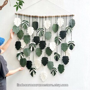 May include: A macrame wall hanging featuring a cascade of green and white leaf shapes. The leaves are made of knotted string and are arranged in a staggered pattern. The wall hanging is suspended from a wooden dowel.