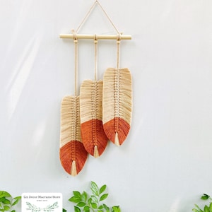 May include: Macrame wall hanging featuring three feather-shaped designs. Each feather has a natural beige upper section and a burnt orange lower section. The feathers hang from a wooden dowel with a natural cord.