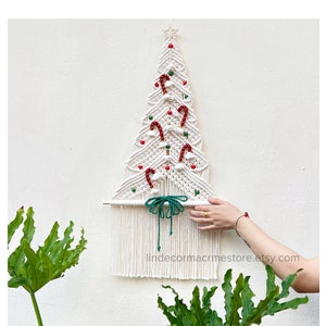Puede incluir: Adorno de pared de árbol de Navidad de macramé blanco con bastones de caramelo rojo y blanco, cuentas verdes y pompones blancos. El árbol está decorado con un lazo verde y una estrella en la parte superior. El texto "lindecormacrmestore.etsy.com" está en la parte inferior de la imagen.